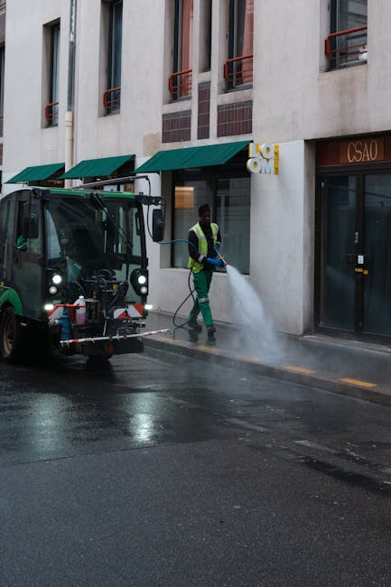 A street-level view of a professional cleaning process on a city pavement, involving a worker in a high-visibility vest operating a street cleaning machine equipped with spray nozzles. The worker is using a hose connected to the machine to clean a concrete sidewalk, which appears wet and freshly cleaned. In the background, there is a multi-storey building with beige walls, red window railings, and green awnings over the storefronts. The scene is lit with natural daylight, highlighting the glossy, cleaned surface of the pavement. This image illustrates a thorough deep cleaning operation, typical of commercial cleaning services provided by West Kensington Cleaners, demonstrating their expertise in surface cleaning and hygiene maintenance.