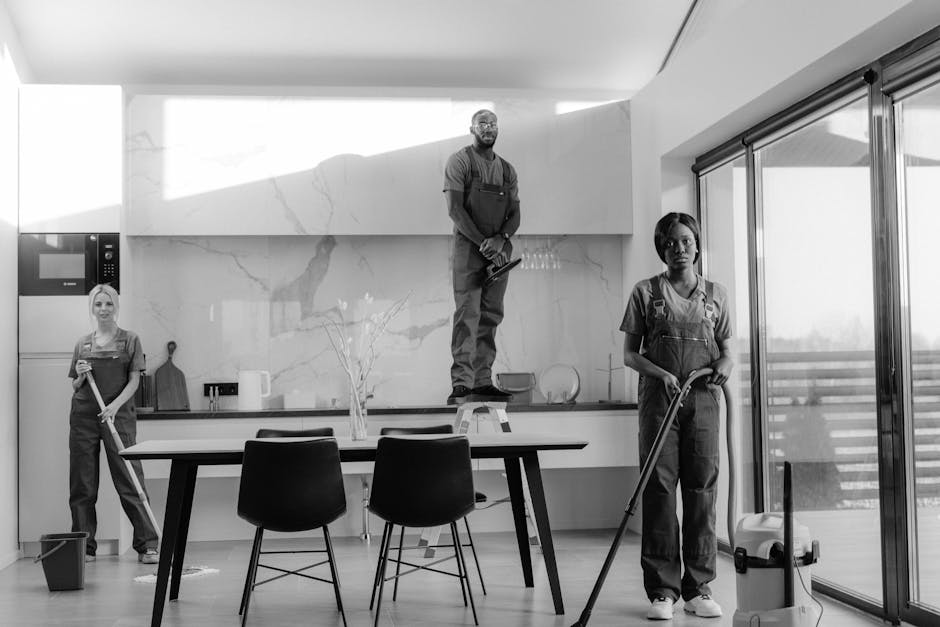 Three professional cleaners from West Kensington Cleaners are performing surface cleaning and sanitisation in a modern kitchen. One cleaner is standing on a small step stool, wiping the upper kitchen cabinets with a cloth. Another is vacuuming the floor near the foreground, while the third is holding a mop and standing on the tiled floor, near a sliding glass door that lets in natural light. The kitchen features a sleek, white marble backsplash, stainless steel appliances, including a microwave oven, and a contemporary dining table with black chairs. The surfaces are visibly clean and shiny, with no dust or debris present, and the room appears well-maintained and hygienic under bright, natural lighting. This scene exemplifies the comprehensive domestic cleaning services provided by West Kensington Cleaners, emphasizing surface cleaning and thorough sanitisation for a pristine home environment.